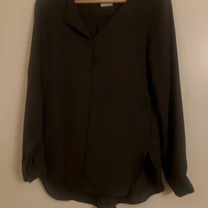 Mörkgrå blus från VILA, strl xs - Snygg mörkgrå blus från VILA i storlek xs. Blusen har v-ringad hals, långa ärmar med knapp vid mudden och slits i sidorna. Materialet känns lätt och mjukt, perfekt för dig som gillar stilrena och enkla plagg.