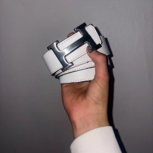 Vitt skärp med silverfärgat H-spänne - Stilrent vitt bälte med ett stort H-format spänne i silverfärg. Bältet har tydliga sömmar längs kanterna och är gjort i ett slätt material som ger en clean look. Perfekt accessoar för att lyfta din outfit.