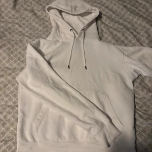 Vit clean hoodie från Jack & Jones - Snygg vit hoodie från Jack & Jones med klassisk huva och snörning. Tröjan har en stor magficka och ribbade muddar vid ärmslut och nederkant. Tillverkad i mjuk bomull som är skön att ha på sig. Perfekt för chill dagar eller när du vill ha en clean look.
