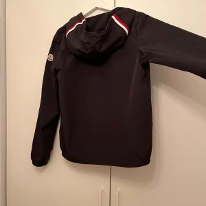 Svart vindjacka från Moncler med huva - Svart vindjacka från Moncler med huva och dragsko, snygga detaljer med röd och vit rand på ryggen. Jackan har dragkedja framtill, två fickor med tryckknappar och Moncler-logga på ärmen. Perfekt nu när det är regnigt. Helt ny.                 pris kan diskuteras