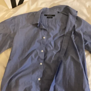 Blå skjorta från Tiger of Sweden - Snygg blå skjorta från Tiger of Sweden med klassisk krage och vita knappar. Skjortan har lång ärm och är tillverkad i bomull, perfekt för en clean och stilren look. Passar dig som gillar enkel och tidlös stil.