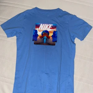 Blå Nike t-shirt med grafiskt tryck - Snygg blå t-shirt från Nike i bomull med ett färgstarkt grafiskt tryck på bröstet. Trycket har futuristiska ansikten, jordklot och solnedgång i starka färger. Klassisk rund hals och korta ärmar. Perfekt för dig som gillar streetwear och coola prints.
