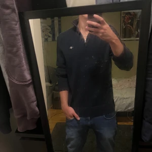 Mörkblå half zip stickad tröja Gant - Snygg mörkblå stickad tröja från Gant med half zip och liten broderad logga på bröstet. Tröjan har hög krage och långa ärmar med ribbade muddar. Perfekt för dig som gillar klassisk stil med en modern touch.