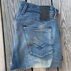 Replay Anbass blå jeans herr - Snygga Replay Anbass jeans i klassisk blå tvätt med slitningar och raka ben. Jeansen har fem fickor, läderpatch bak och coola detaljer på bakfickan. Perfekt passform och stilren look för dig som gillar street och casual.