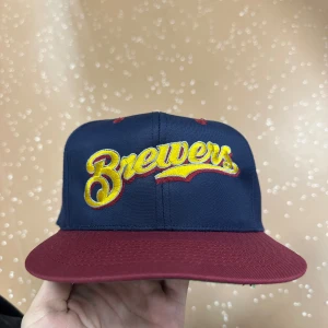 Brewers snapback keps marinblå vinröd - Snygg snapback keps från Brewers med marinblå krona och vinröd skärm. Broderad gul och röd Brewers-logga framtill, grönt underskärm och justerbar baksida. Perfekt för dig som vill ha sportig stil med lite retrovibe.