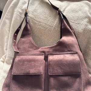 Brun mocka axelväska från Cicelle - Snygg brun axelväska i mocka från Cicelle med två stora fickor framtill och guldfärgade detaljer. Justerbar axelrem med metallspänne och nitar. Väskan har magnetknappar och ett rymligt innerfack med dragkedja. Perfekt för dig som gillar stilrena accessoarer.