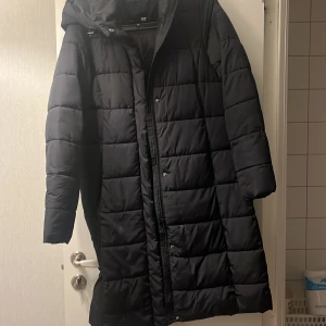 Svart lång pufferjacka med huva - Säljer en svart lång pufferjacka från Lager 157 med huva och quiltad design. Jackan har dragkedja och tryckknappar framtill, samt långa ärmar. Väldigt bra skick, använt ungefär 5 gånger! Köparen står för frakten (69-79)kr 