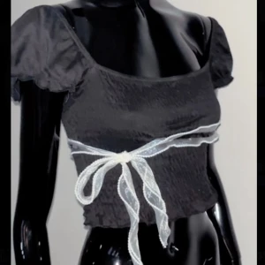 Svart croppad topp med rosett - Supersöt svart croppad topp med puffiga korta ärmar och en transparent vit rosett som knyts runt midjan. Toppen har en smockad struktur som ger en snygg och figurnära passform. Perfekt för dig som gillar trendiga detaljer och romantisk vibe.