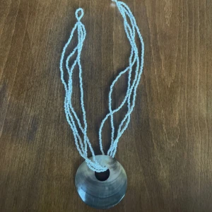 Halsband med pärlor och stor rund berlock - Unikt halsband med flera tunna vita pärlsträngar och en stor, rund gråskimrande berlock i mitten. Berlocken har en slät, glansig yta och ett hål i mitten för en cool statement-look. Perfekt för dig som gillar att sticka ut med accessoarer.