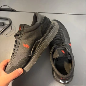 Svarta Polo Ralph Lauren sneakers - Svarta sneakers från Polo Ralph Lauren med röd broderad logga på sidan och hälen. Skorna har snörning, platt sula och rund tå. Materialmix av skinn, textil och syntet. Snygg kontrast mellan svart och rött, perfekt för dig som gillar klassisk streetstil.