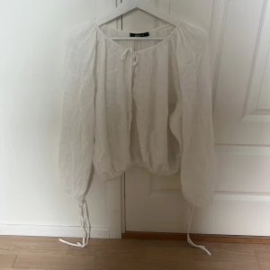 Vit blus med snörning Gina Tricot - Luftig vit blus från Gina Tricot i linne med långa ärmar och snörning både vid halsringningen och ärmsluten. Skjortan har en avslappnad passform och en lätt skrynklig textur som ger en chill vibe.