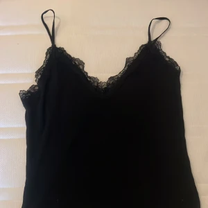 Svart spetskantad linnetopp från Amisu - Svart linnetopp från Amisu med tunna axelband och v-ringad hals. Toppen har snygga spetsdetaljer längs halsringningen och en lätt vågig kant nedtill. Materialet är mjukt och består av viskos och elastan, vilket gör den skön och följsam.