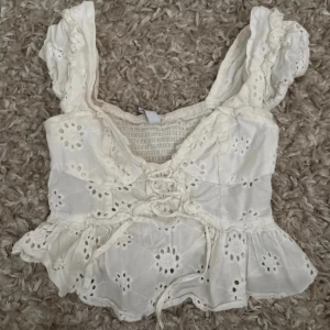 Vit broderad blus med snörning - Säljer en söt vit blus med broderade blommor och snörning framtill. Toppen har volangdetaljer vid axlar och nederkant, samt breda axelband. Perfekt för sommarens alla tillfällen, superfin till jeans eller kjol!