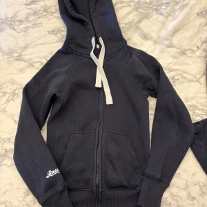 Russedress sett  - Mörkblå hoodie och mjukisbyxor från Russedress med vit snörning i båda plaggen. Tröjan har känguruficka och tryckt logga på ärmen. Tillverkad i mjuk bomullsmix, perfekt för chill dagar. Långärmad och med ribbade muddar för extra komfort.