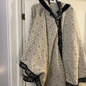 Grå och svart stickad poncho från Norge  - Säljer en mysig stickad poncho från norge i grått och svart med traditionellt mönster och snygga metallspännen framtill. Jackan har huva och breda ärmar, samt dekorativa kanter i svart med grått mönster. Perfekt för dig som gillar unik stil och nordisk vibe.