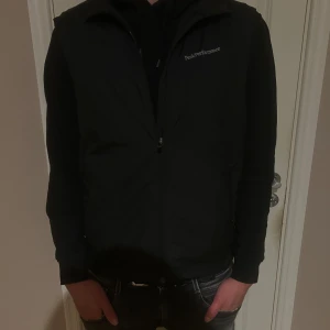 Svart dunväst Peak Performance L - Svart dunväst från Peak Performance i storlek L. Västen har en stilren design med diskret logga på bröstet och dragkedja framtill. Perfekt att slänga över en hoodie för extra värme. Materialet är syntet och dun, med två sidofickor med dragkedja.