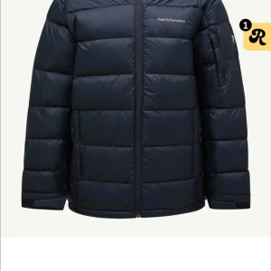 Svart dunjacka Peak Performance - Snygg svart dunjacka från Peak Performance med quiltad design och dragkedja framtill. Jackan har lång ärm och diskret logga på bröstet. Perfekt för kalla dagar när du vill hålla värmen och samtidigt se fräsch ut.