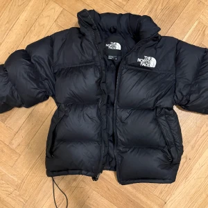 Svart pufferjacka från The North Face - Säljer en svart pufferjacka från The North Face med broderad logga på både bröstet och ryggen. Jackan har dragkedja  och är långärmad. Har även luva om man vill ta ut den. Perfekt för kalla dagar med sin tjocka och fluffiga design. 
