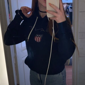 Mörkblå hoodie från Gant - Säljer en mörkblå hoodie från Gant med vit snörning och tryckt logga framtill. Tröjan har huva och en klassisk collegekänsla, perfekt till jeans eller joggers. Skön och avslappnad passform.