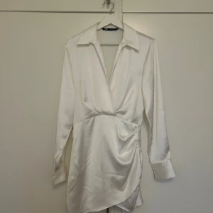 Vit omlottklänning i satin från Zara - Supersnygg vit kort omlottklänning från Zara i glansig satin. Klänningen har lång ärm,  krage, markerad midja och draperad detalj framtill. Dold dragkedja i sidan för en smidig passform. Användes som min studentklänning och använd endast en gång.