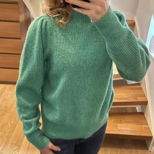 Grön stickad tröja från Neo Noir - Mysig grön stickad tröja från Neo Noir med ribbad struktur och lätt puff vid axlarna. Tröjan har hög hals och långa ärmar, perfekt för lager på lager under kyliga dagar.