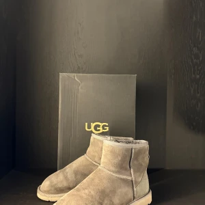 Grå UGG boots i mocka - Säljer ett par klassiska UGG boots i grå mocka med rund tå och platt sula. LÅDA KOMMER MED. Skorna har en mjuk och fluffig insida som håller fötterna varma under kyliga dagar. Perfekt för dig som gillar en chill och cozy stil. UGG-logga på hälen och grov yttersula för extra grepp.