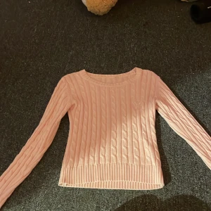 Ljusrosa kabelstickad tröja - Supersöt ljusrosa kabelstickad tröja med rund halsringning och långa ärmar. Tröjan har ribbade muddar vid ärmslut och nederkant, vilket ger en snygg och klassisk look. Perfekt för lager-på-lager under kyligare dagar. Vet varken märke eller storlek men skulle säga att den är storlek S, det står CLFD PÅ BRÖSTET 