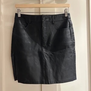 Svart kortkjol i skinn från Noisy May - Svart kortkjol från Noisy May i skinnimitation med klassisk femficksdesign, knapp och dragkedja framtill samt bälteshällor. Kjolen har en rak passform och är perfekt för dig som gillar en edgy och stilren look.