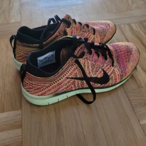 Nike Free Flyknit sneakers färgglada - Nike Free Flyknit sneakers med stickad ovandel i mixade färger som rosa, orange och gul. Svarta detaljer och snörning, samt en ljusgrön platt sula med hexagonmönster undertill. Lätta och flexibla med rund tå, perfekt för träning eller chill.