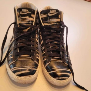 Nike Blazer Highs med svartvitt mönster - Säljer ett par Nike Blazer Highs med coolt svartvitt randigt mönster. Skorna har snörning, rund tå och platt sula. Ovandelen är i skinn med kontrasterande vita och svarta detaljer, samt tydlig Nike-logga på plösen och hälen. Perfekt för dig som vill sticka ut med dina sneakers.