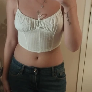 Vit broderad korsett-topp med knyt - Superfin vit korsett-topp med tunna axelband och broderade detaljer upptill. Toppen är croppad, har knyt framtill och dragkedja i ryggen. Materialet känns som bomull och har diskreta ränder. Perfekt för sommarens alla tillfällen!