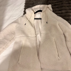Vit teddyjacka med huva från H&M - Mysig vit teddyjacka från H&M med huva och snörning. Perfekt för kalla dagar och har en fluffig, mjuk känsla. Passformen är normal och är i storlek S men passar även M. Jackan är i mycket bra skick!