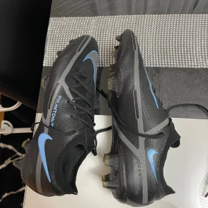 Nike Phantom GT2 Elite fotbollsskor svart/blå - Nike Phantom GT2 fotbollsskor i svart med blå och grå detaljer. Snygg, modern design med snörning och platt sula för grym kontroll på planen. Ovandelen är tillverkad i syntetmaterial med textur för bättre bollkänsla. Perfekt för dig som vill sticka ut på fotbollsplanen. 