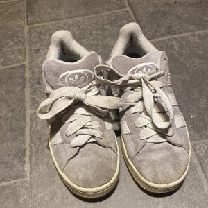 Grå Adidas Campus sneakers mocka - Säljer ett par grå Adidas Campus sneakers med klassiska vita ränder och logga på plösen och hälen. Skorna har rund tå, platt sula och är tillverkade i mjuk mocka med vita detaljer i skinn. Perfekta för dig som gillar retrostil och streetwear.