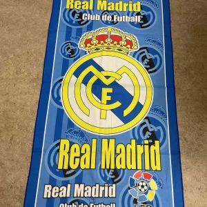 Real Madrid halsduk med klubbmärke - Snygg halsduk med Real Madrid klubbmärke och LFP-logga. Färgerna är blå, gul, vit och inslag av röd. Halsduken har ett randigt mönster i bakgrunden och stora tryckta loggor. Perfekt för dig som älskar fotboll och vill visa ditt stöd för laget.