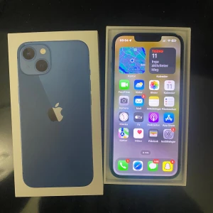 iPhone 13 128GB Blue - Snygg blå iPhone 13 med 128GB lagring. Telefonen levereras i originalförpackning. Skärmen och baksidan ser fina ut, men det finns en spricka i hörnet på baksidan. Kraftfull kamera, Face ID och lång batteritid. Perfekt för dig som vill ha en modern och pålitlig smartphone.