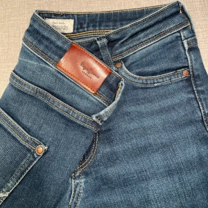 Blå jeans från Pepe Jeans London - Snygga blå jeans från Pepe Jeans London med klassisk femficksdesign och slitna detaljer vid fickan. Jeansen har kontrastsömmar och en brun läderpatch bak i midjan. Modellen har en rak passform och är tillverkad i mjukt denimtyg.