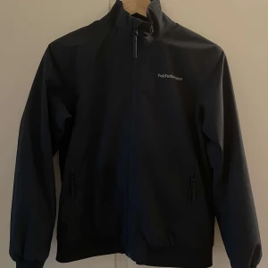 Mörkblå vindjacka från Peak Performance  - Mörkblå vindjacka från Peak Performance med stilren design och broderad logga på bröstet. Jackan har hög krage, dragkedja framtill och två fickor med dragkedja. Materialet är slätt och syntetiskt, perfekt för blåsiga dagar.