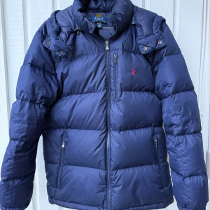 Mörkblå pufferjacka Polo Ralph Lauren Junior - Säljer en mörkblå pufferjacka från Polo Ralph Lauren med avtagbar huva, dragkedja och fickor med zip. Jackan har det klassiska röda Polo-logot på bröstet. Lite små repor på baksida vänster ärm som lagats annars väldigt fint skick! Använd två säsonger av 15-16 åring. 