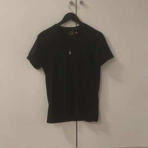 Svart t-shirt Polo Ralph Lauren - Svart t-shirt från Polo Ralph Lauren med klassisk rund hals och vit broderad logga på bröstet. Modellen är slim fit och har korta ärmar. Materialet är mjuk bomull som känns skönt mot huden. Perfekt för dig som gillar stilrena och tidlösa plagg.