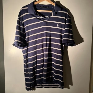 Randig marinblå pikétröja Polo Ralph Lauren - Snygg marinblå pikétröja från Polo Ralph Lauren med vita horisontella ränder och klassisk krage. Tröjan har korta ärmar och den ikoniska broderade loggan på bröstet. Tillverkad i mjuk bomull, perfekt för en chill och stilren look.