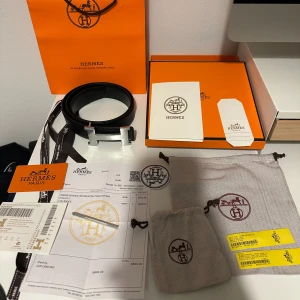 Svart Hermès bälte med H-spänne - Säljer ett klassiskt svart bälte från Hermès med ikoniskt H-spänne i silverfärgad metall. Bältet är gjort i exklusivt skinn med en stilren och tidlös design. Kommer med originalbox, dustbags, kvitto och certifikat. Perfekt accessoar för att lyfta vilken outfit som helst.