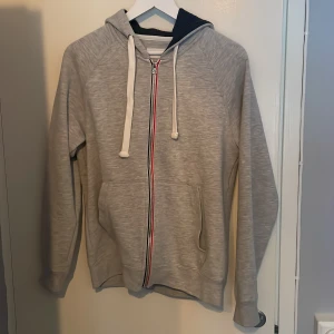 Grå hoodie  - Grå hoodie från Pier One med vit snörning och hel dragkedja med röd och mörkblå detalj. Tröjan har en mörkblå insida på huvan. Använd 1 gång så i väldigt fint skick. Storlek XS men passar även S. 