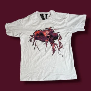 Vit Vlone t-shirt med grafiskt tryck - Vit t-shirt från Vlone i 100% bomull med ett stort grafiskt tryck på bröstet som föreställer ett ansikte med lila och röda toner, samt en stor V på ryggen med blixtliknande detaljer. Klassisk rund hals och korta ärmar. Perfekt statement-plagg för streetwear.