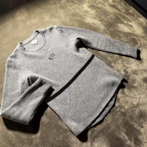 Grå stickad tröja från GANT Rugger - Ribbstickad grå tröja från GANT Rugger med svart broderad logga på bröstet och en svart horisontell rand över magen. Rund halsringning och långa ärmar. Perfekt för kyliga dagar och enkel att matcha med jeans👖