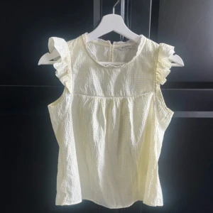 Randig blus med volang från Stradivarius - Supersöt blus från Stradivarius i storlek S, med ljusgula och vita ränder. Blusen har volangdetaljer vid axlarna, är ärmlös och har spetskant vid halsen. Materialet är lätt och bomulligt med en skön struktur. 