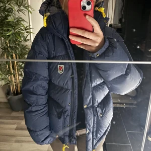 Mörkblå dunjacka från Fjällräven - Säljer en mörkblå dunjacka från Fjällräven med gul snörning i huvan och nederkant. Jackan har två stora fickor med knappar, huva och Fjällrävens klassiska logga på bröstet. Använd 1 vinter. Inga defekter 