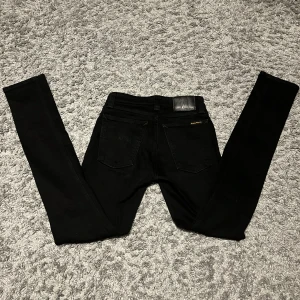 Svarta jeans från Nudie Jeans - Säljer ett par svarta skinny jeans från Nudie Jeans i storlek W26 L32. Jeansen har klassisk femficksdesign, svarta sömmar och Nudie Jeans logga på bakfickan samt läderpatch i midjan. Snygg passform med smala ben och stretchigt jeanstyg.
