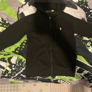 Svart hoodie från Polo Ralph Lauren - Svart hoodie från Polo Ralph Lauren med dragkedja framtill, huva med snörning och klassisk vit logga på bröstet. Tröjan har känguruficka och är långärmad. Perfekt för en chill och stilren look.äkta