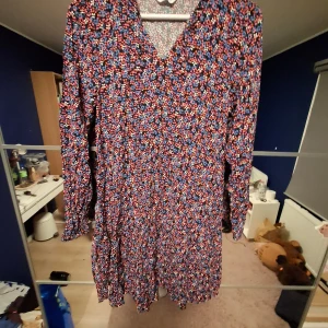 Blommig kortklänning med v-ringning - Säljer en färgglad kortklänning med blommigt mönster i blått, rosa, rött och vitt. Klänningen har v-ringning, långärmade ärmar och ett flowigt fall. Perfekt för dig som gillar mönstrade plagg med mycket färg och vill sticka ut lite extra.